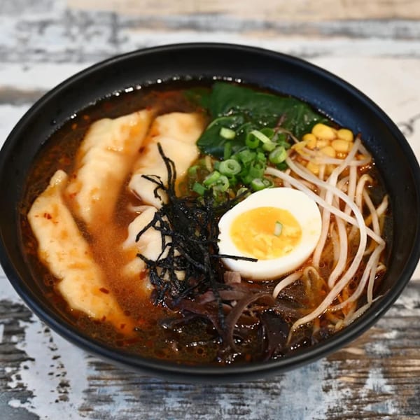 A black bowl of ramen with dumplings, a soft-boiled egg, corn, scallions, and seaweed.