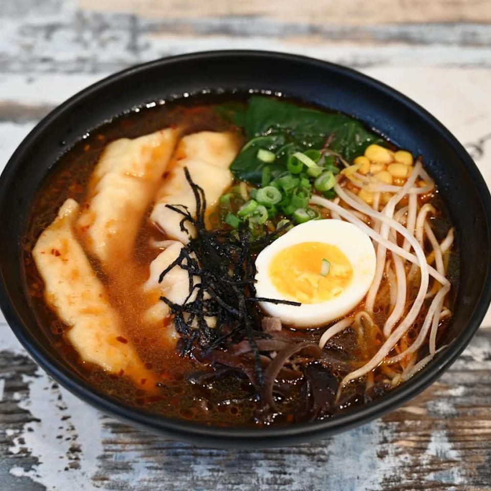 A black bowl of ramen with dumplings, a soft-boiled egg, corn, scallions, and seaweed.
