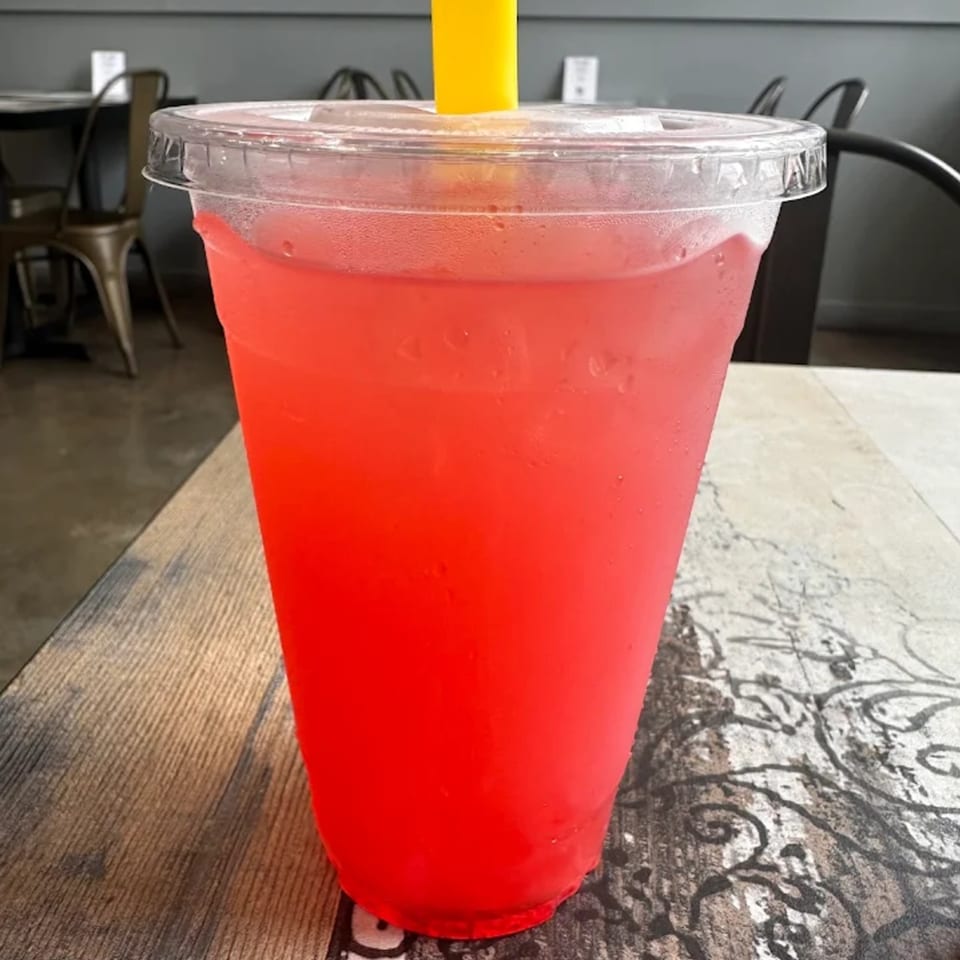 A vibrant red drink in a clear plastic cup with a yellow straw sits on a patterned wooden table.