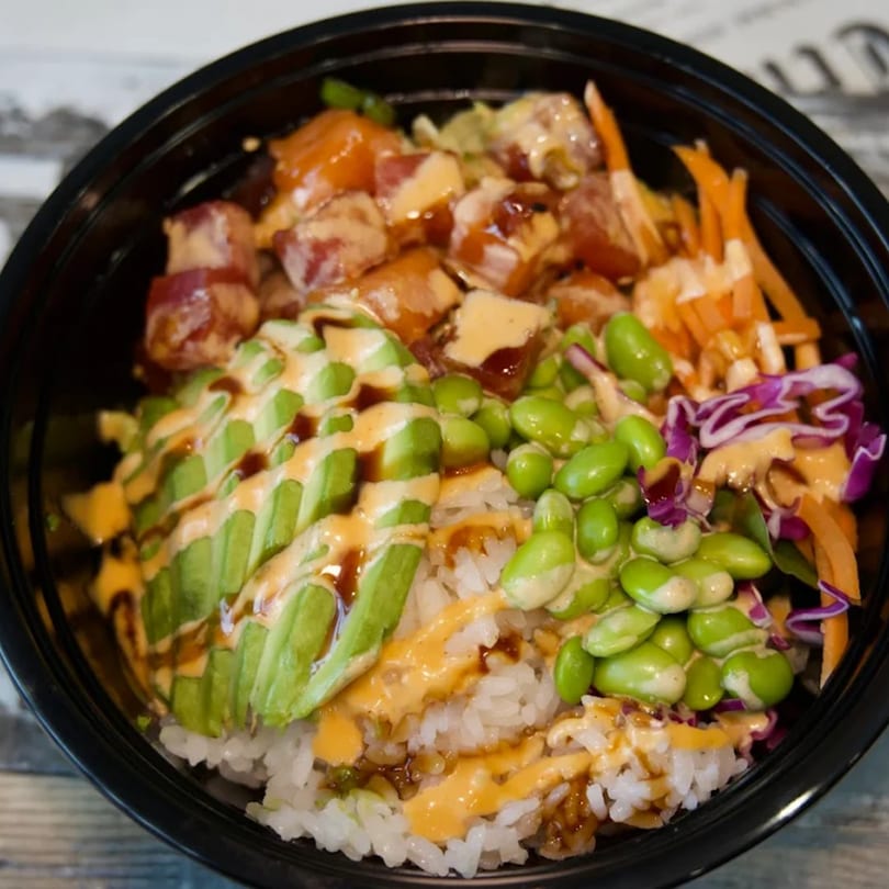 A colorful poke bowl with tuna, avocado, edamame, carrots, and purple cabbage over rice.